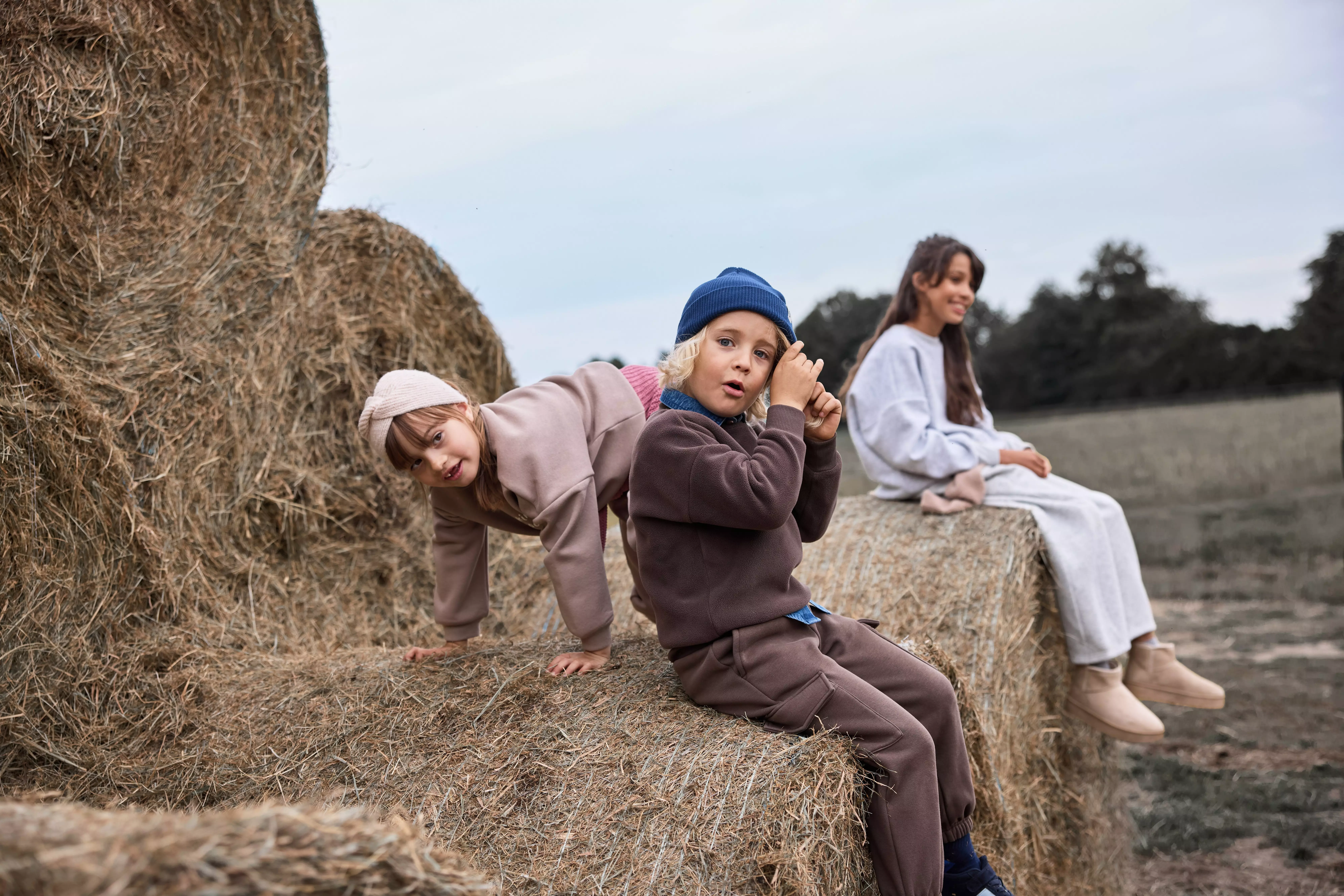 Pepco children and woman wearing casual outfits, sitting and playing on stacked hay bales outdoors.